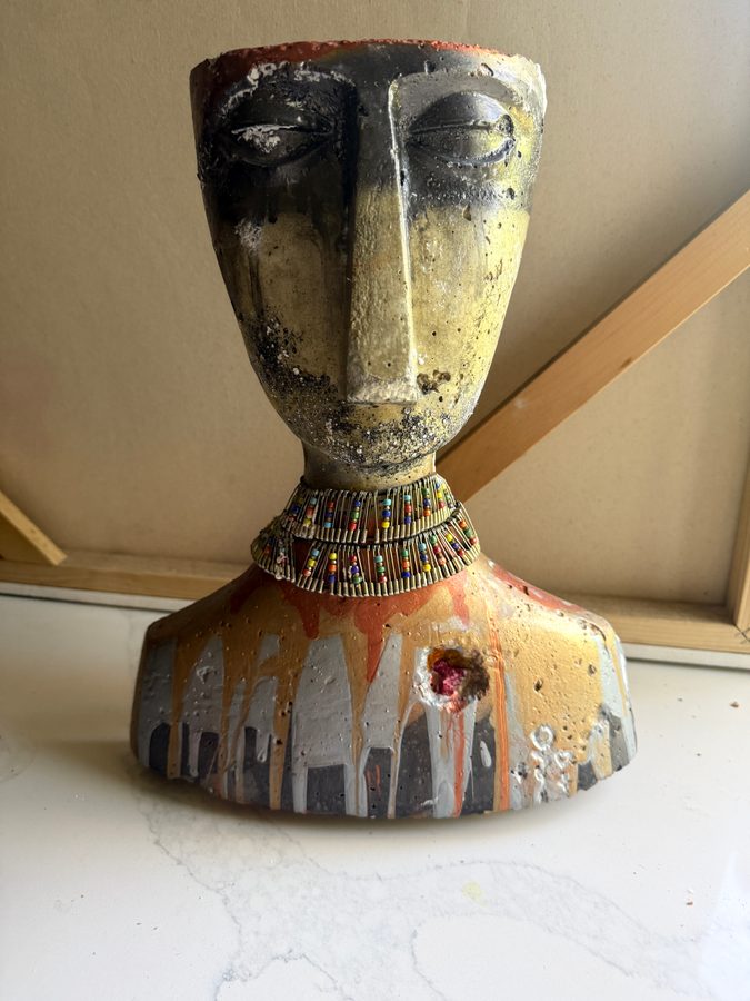 The Warrior: a hand-sculpted cement bust with a long face and closed eyes, weathered black and gold, wearing a beaded collar